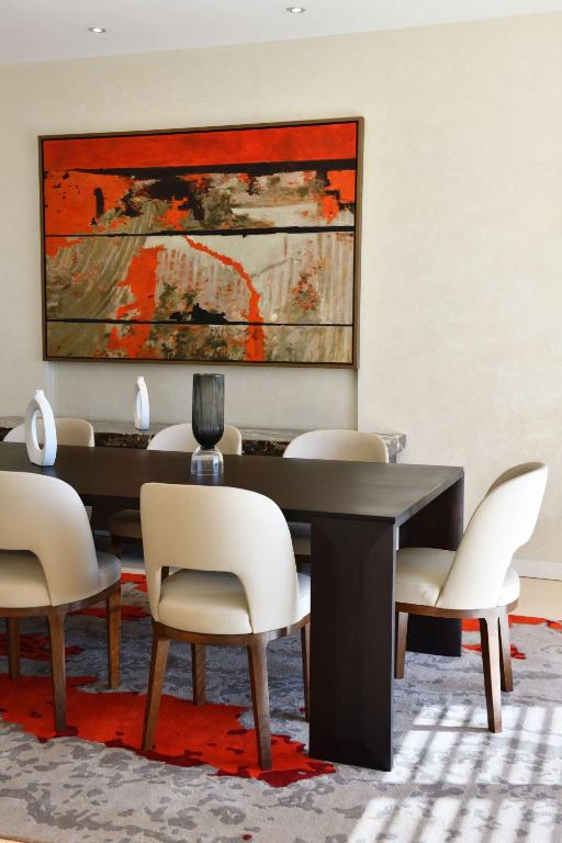 Mūsdienīgs ēdamistabas interjers ar tumšu galdu, baltām krēsliem un krāsainu mākslas darbu uz sienas. Uz grīdas ir stilīgs paklājs ar sarkanu un pelēku dizainu.