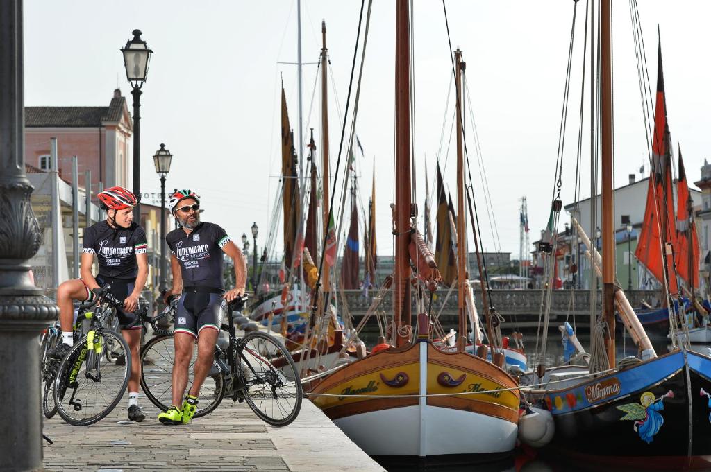 Divi riteņbraucēji atpūšas pie krasta, blakus krāsainām laivām un vēsturiskām ēkām. Fons ir gleznains, ar skaidru debesu un ostas atmosfēru.