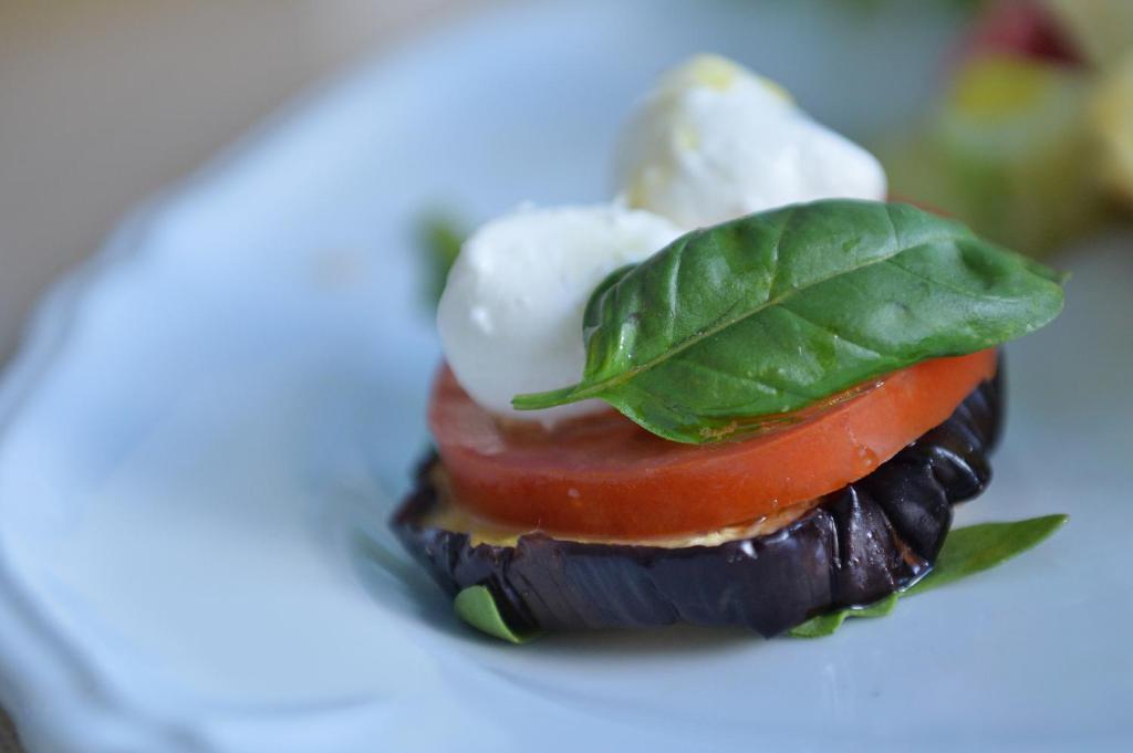 Miniatūrā dārzeņu uzkoda ar baklažānu, tomātu, mocarellu un baziliku, pasniegta uz balta šķīvja.
