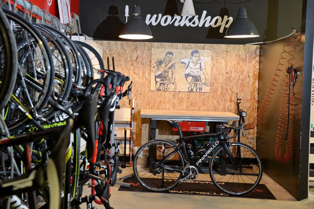 Velosipēdu darbnīca ar velosipēdu stendu un rīku galdu, sienā ir glezna ar riteņbraucējiem. Gaismas piekaramie lampas rada siltu atmosfēru.