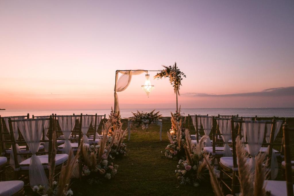 Romantiska kāzu ceremonijas vieta pie jūras, apgaismotā ar lustras gaismu un dekorēta ar ziediem un staltajiem zālēm. Saule riet un rada maigu atmosfēru.