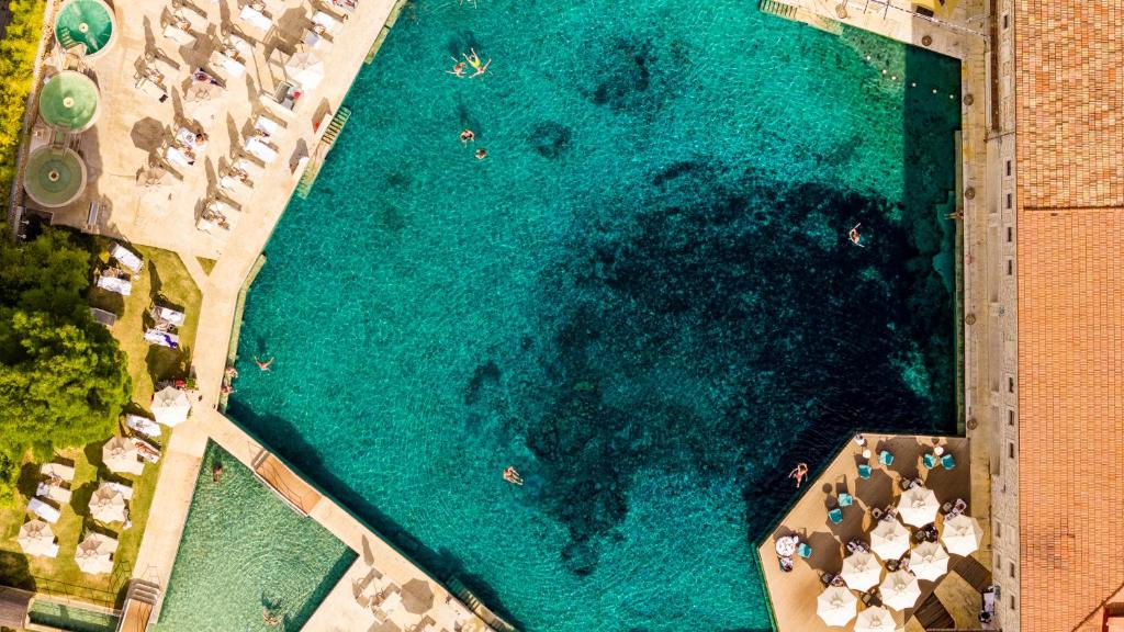 Lidošanas baseins ar dzidriem ūdeņiem un sauļošanās krēsliem apkārt, daži cilvēki peld un atpūšas. Skats no augšas uz relaksējošu atpūtas vidi.