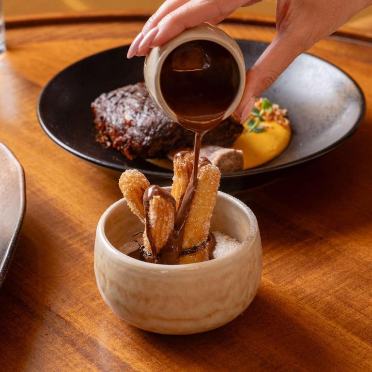 Churros tiek pasniegti ar šokolādes mērci, kas tiek lejama virs tiem. Fons ir tumšs deserts un krēmīga mērce, kas rada patīkamu gaisotni.