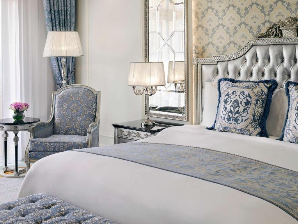 Elegants guļamistabas interjers ar sudraba krāsas gultu, greznām spilvendrānām un zilu aksesuāru. Telpā ir arī stilīgs krēsls un dekoratīvs galdiņš ar ziediem.