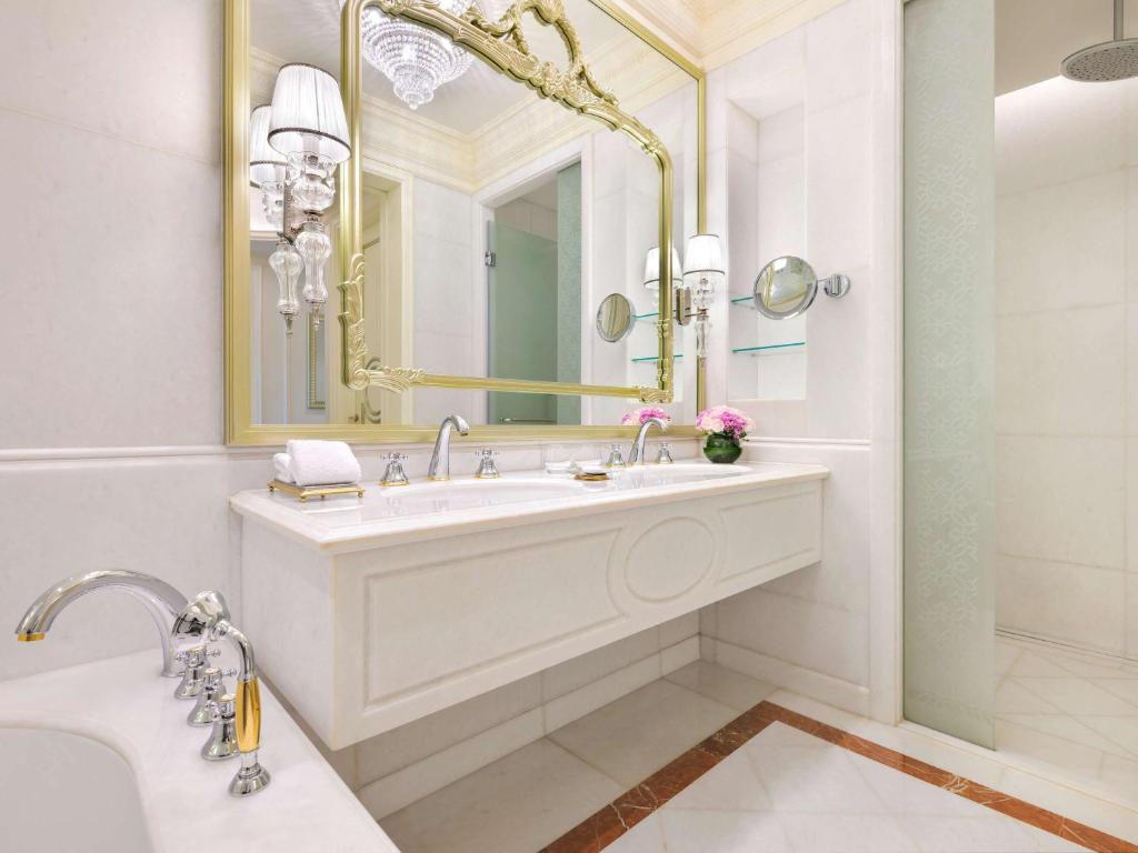 Elegants vannas istabas interjers ar baltiem akmens apdarēm, greznu spoguli ar zelta rāmīti un mūsdienīgu dušas kabīni. Vannas istabā ir arī stilīgas apgaismojuma ķermeņi un dekoratīvas puķes.