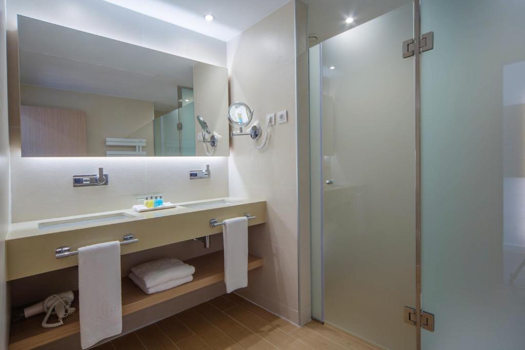 Mūsdienīgs vannas istabas interjers ar plašu spoguli, divām izlietnēm un stiklota dušas kabīni. Uz galda redzami higiēnas piederumi un dvieļi.