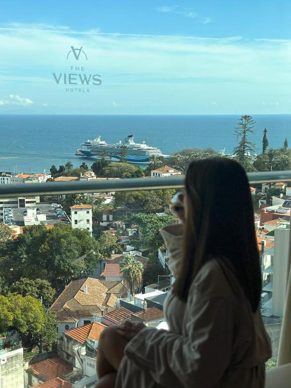 Skats no viesnīcas loga uz ostu, kur redzami kruīza kuģi un pilsētas ainava ar sarkaniem jumtiem un zaļām kokos. Viesis, ģērbtā halātā, bauda mierīgu mirkli.