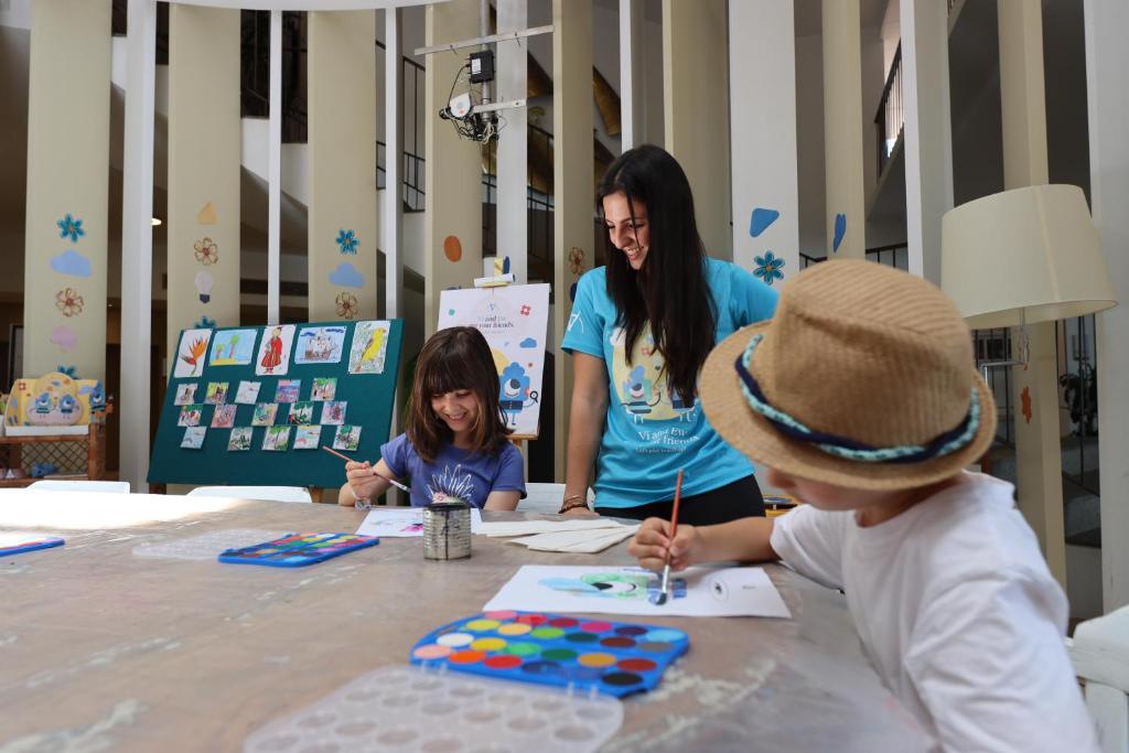 Bērni radoši nodarbojas ar zīmēšanu un krāsošanu, kamēr pieaugušais viņiem palīdz un uzrauga procesu. Interesanta un radoša vide ar mākslas darbiem fona.