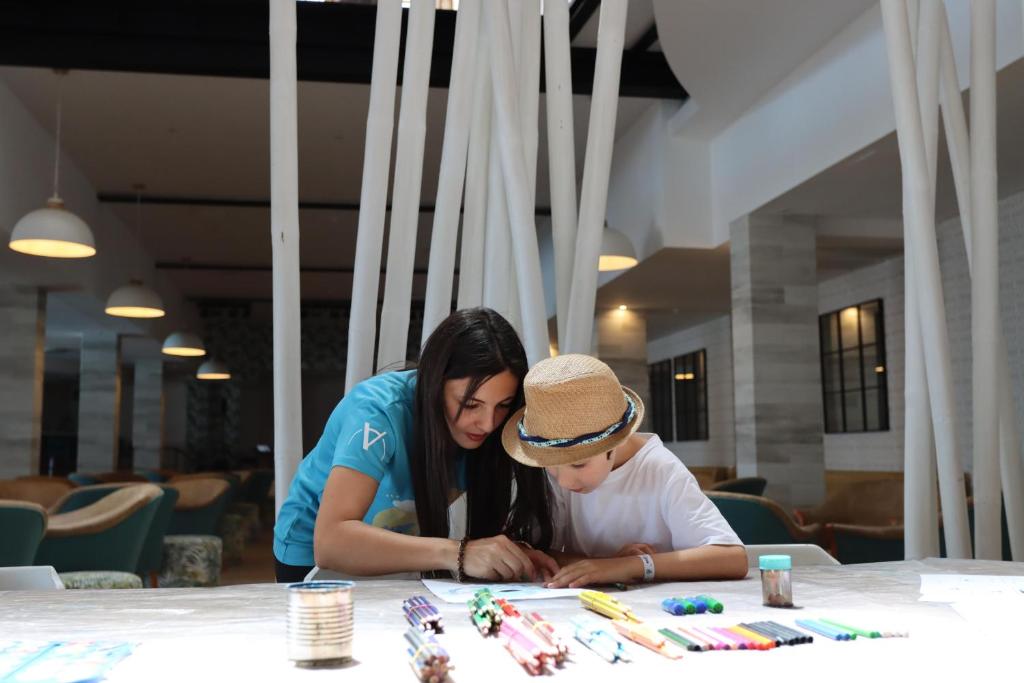 Bērns un pieaugušais kopā zīmē un mākslinieciski darbojas, izmantojot krāsainus marķierus un zīmuļus. Fons ir moderns, ar gaišām gaismām un koka elementiem.