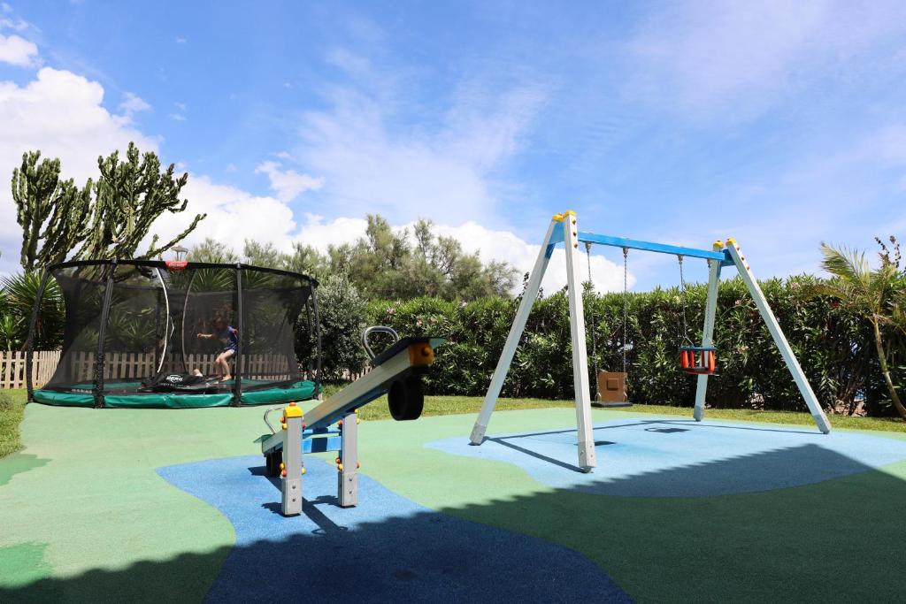 Bērnu rotaļu laukums ar batutu un šūpolēm, zaļā zālē, zem zila debesu fona. Rotaļu laukums ir aprīkots ar krāsainu segumu un privātu žogu.