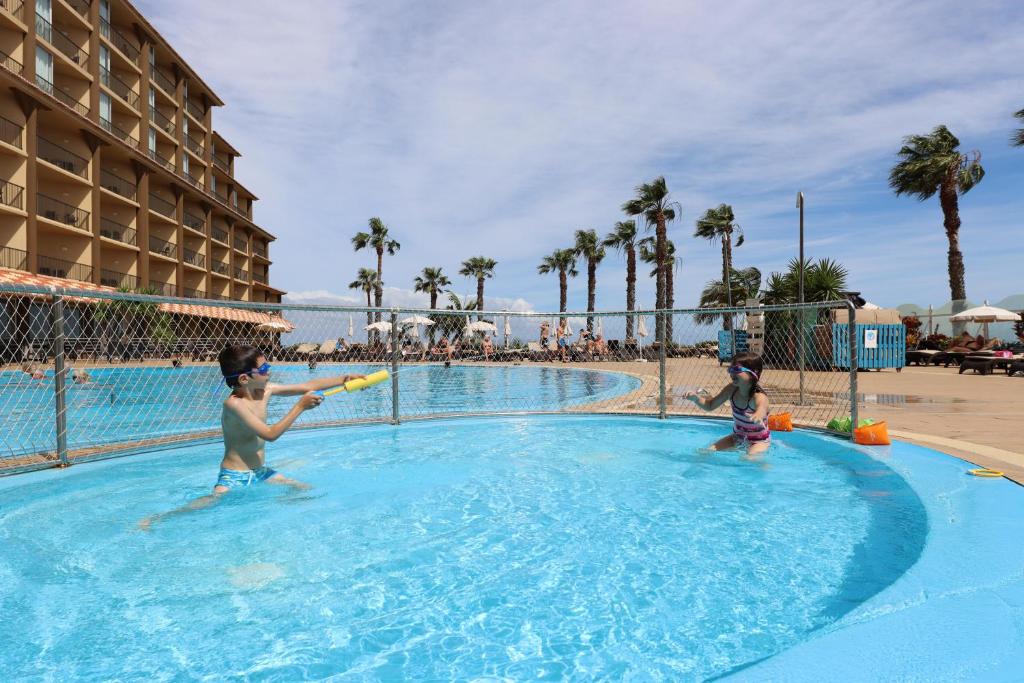Bērni spēlējas baseinā, izmantojot ūdens pistoles, kamēr fonā redzams viesnīcas ēka un palmām apstādīta teritorija. Saulaina diena ar skaidru debesu fonu.