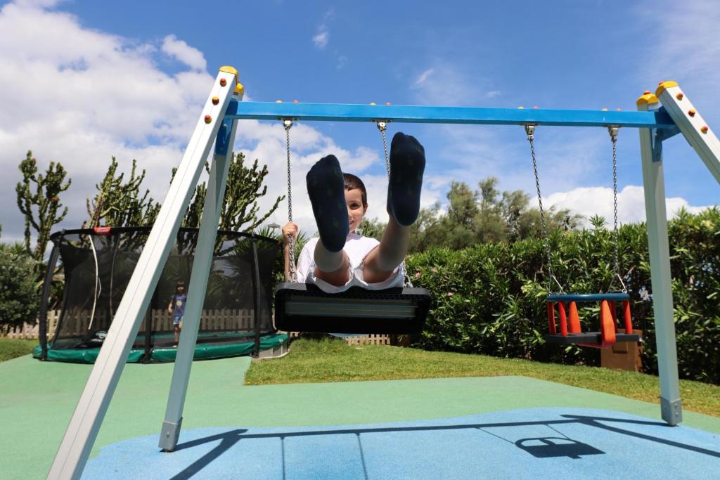 Bērns šūpojas bērnu laukumā ar zaļo zāli un zilu šūpoles rāmi, fonā redzams batuts un krūmi.