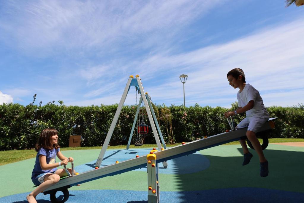 Bērni spēlējas bērnu laukumā uz šūpolēm, baudot saulainu dienu. Fonā redzams zils debesis un zaļš krūmājs.