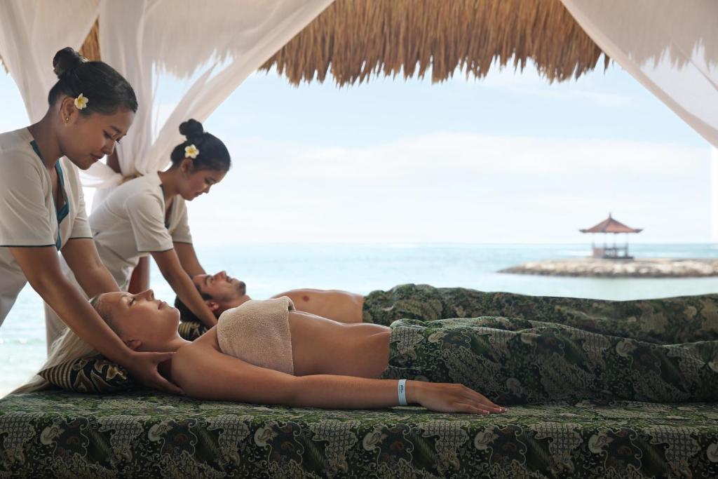 Atpūtas brīdis spa centrā pie pludmales, kur divas masieres sniedz masāžu divām atpūtnieču grupām. Daba un mierīga atmosfēra veido relaksējošu pieredzi.