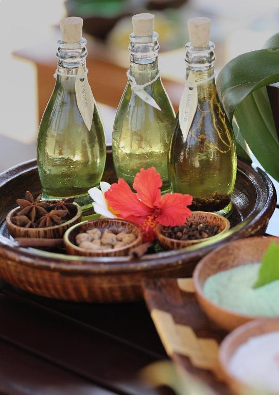 Trīs stikla pudeles ar zaļu eļļu atrodas uz koka šķīvja, blakus ziediem un garšvielām. Šis attēls atspoguļo dabīgu skaistumkopšanas produktu un aromterapijas sastāvdaļas.