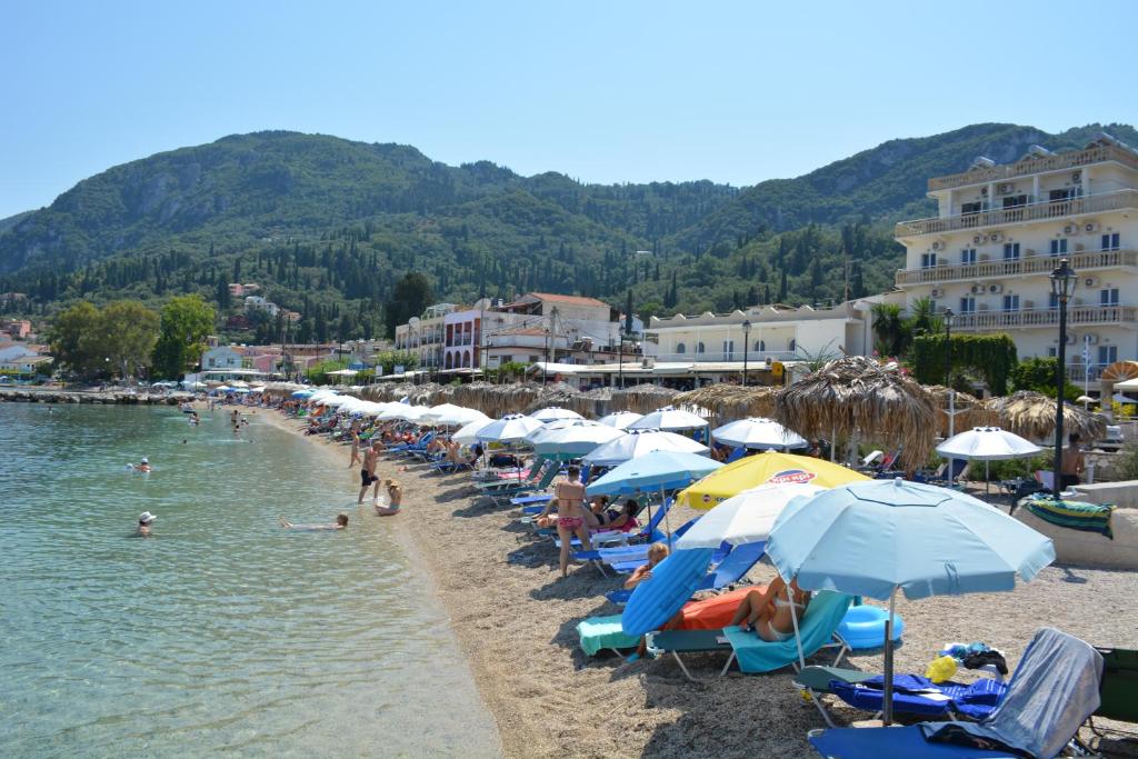 Pludmales ainava ar saulainu laiku, kurā cilvēki atpūšas zem saulessargiem un peldas kristāldzidrajā ūdenī. Fona kalni un viesnīcas papildina relaksējošo atmosfēru.