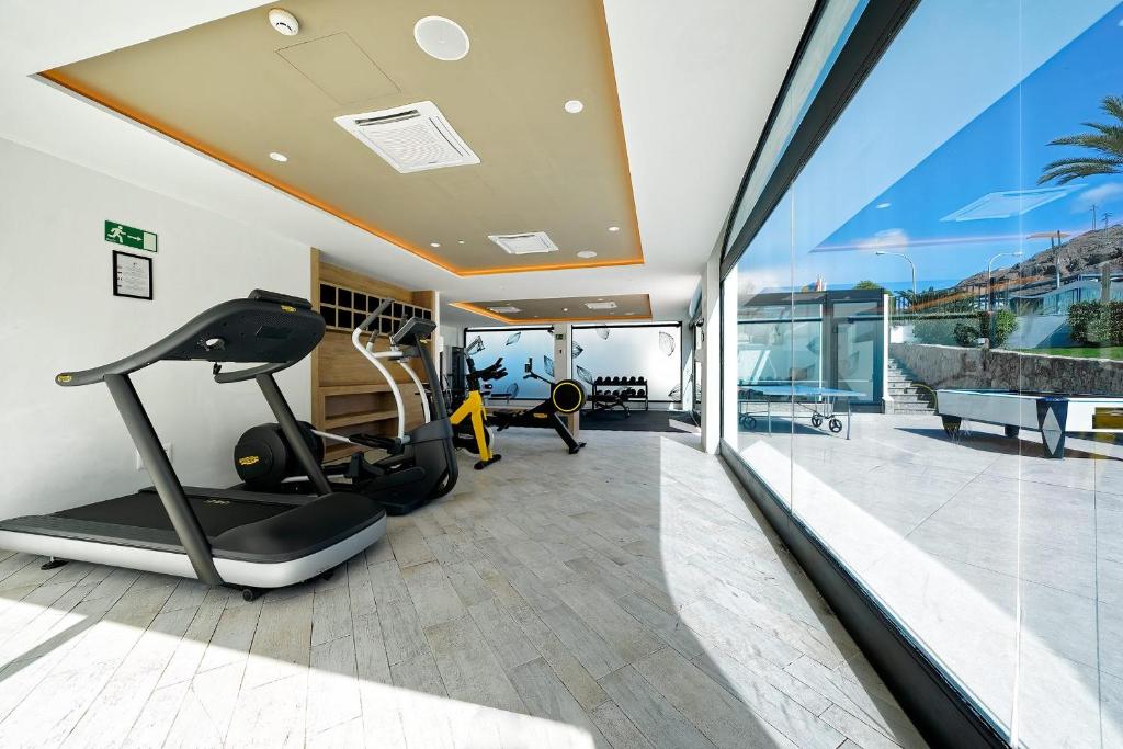Mūsdienīgs sporta zāles interjers ar dažādām fitnesa iekārtām un lieliem logiem, kas nodrošina dabisko apgaismojumu.