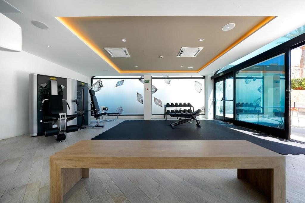 Mūsdienīgs sporta zāles interjers ar trenažieriem un svaru stendu, apgaismots ar siltu apgaismojumu un stikla sienām, kas rada atvērtu un gaisīgu atmosfēru.