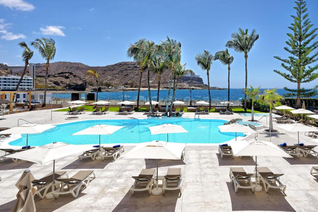 Peldbaseins ar palmām un sauļošanās krēsliem, kas atrodas pie ūdens, ar kalnu ainavu fonā, ideāla vieta relaksācijai un atpūtai.