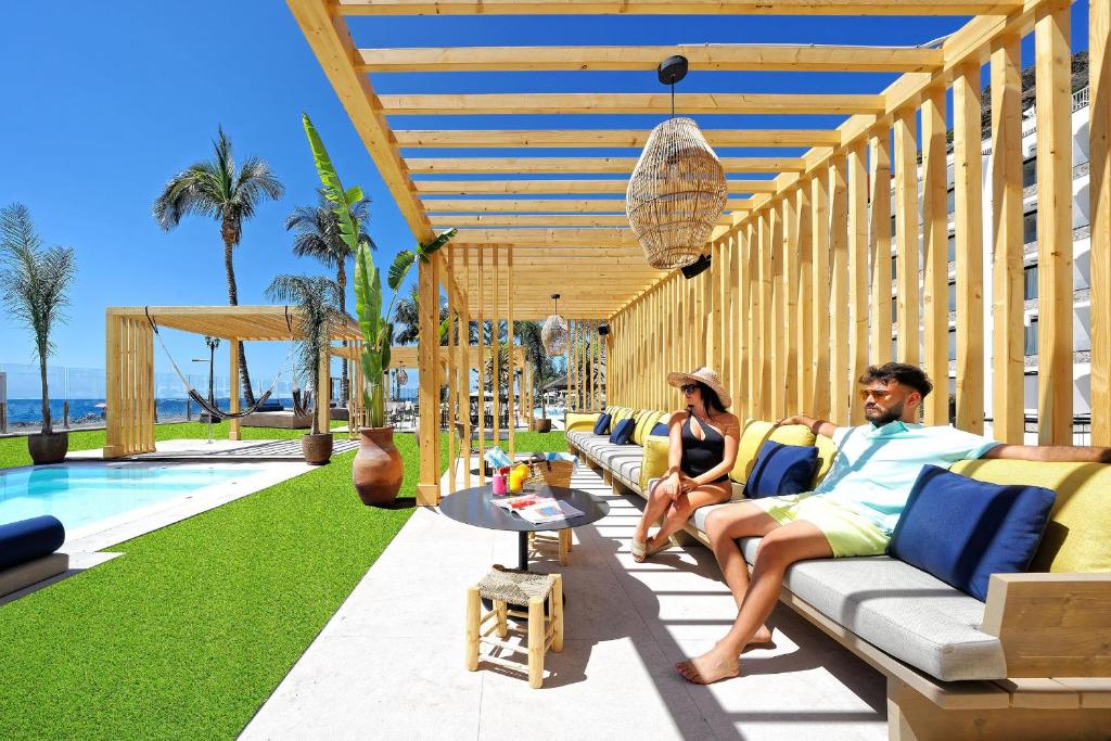 Relaksējoša atpūtas zona pie baseina ar koka nojumi, ērtām mēbelēm un zaļu zālienu, kurā divi cilvēki bauda sauli un skatu uz jūru.