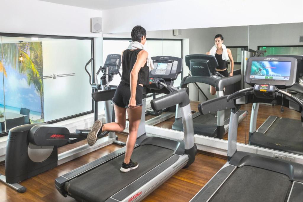 Sieviete vingro uz skrejceliņa sporta zālē ar modernām iekārtām un atspoguļojumu spogulī. Fons ietver pludmales ainavu caur logu, radot relaksējošu atmosfēru.