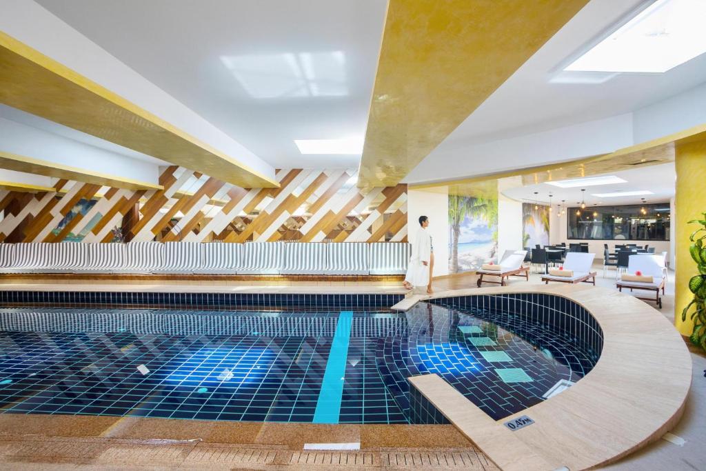 Luksusa iekštelpu peldbaseins ar mūsdienīgu dizainu un relaksējošu atmosfēru, kurā redzams cilvēks baltā apģērbā. Elegantā interjers ar koka akcentiem un gaišām krāsām rada mierīgu noskaņu.