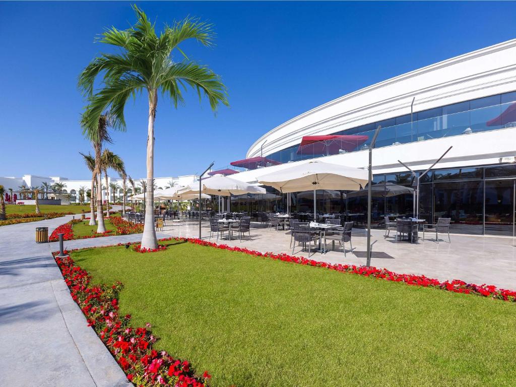 Moderns restorāns ar āra terasi un ziedu ainavu, kur var baudīt ēdienus saulainā vidē. Apkārt ir zaļa zāliena un palmām rotāta teritorija.