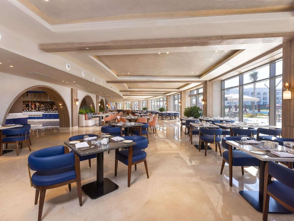 Mūsdienīgs restorāns ar plašām logiem un elegantu interjeru, kurā ir zili krēsli un stilīgi galdi. Telpā ir redzams bārs un zaļi augi, kas piešķir atmosfēru.