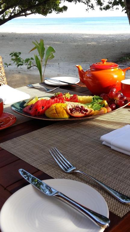 Krāšņs augļu plates ar arbūziem, mango, granātāboliem un vīnogām, kas novietota pie pludmales ar zilu jūru fona, blakus sarkanai tējas kanna un krūzēm.