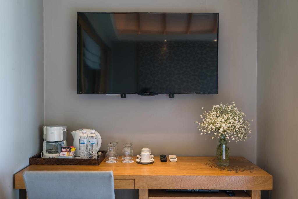 Mūsdienīgs viesnīcas numura stūrī redzams televizors virs koka galda, uz kura atrodas kafijas automāts, ūdens pudeles un ziedu pušķis.