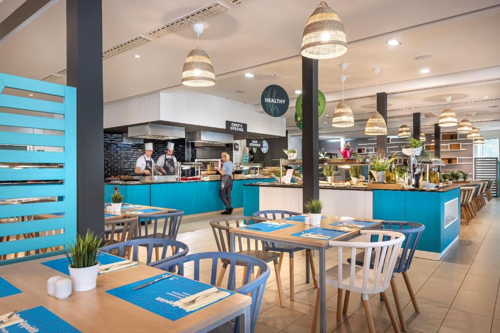 Mūsdienīgs restorāna interjers ar zilu un baltu krāsu shēmu, kurā redzami pavāri, kas strādā virtuvē, un viesējo ēdināšanu. Galdi ir klāti ar ziliem galdautiem un krēsli dažādās krāsās, radot patīkamu atmosfēru.