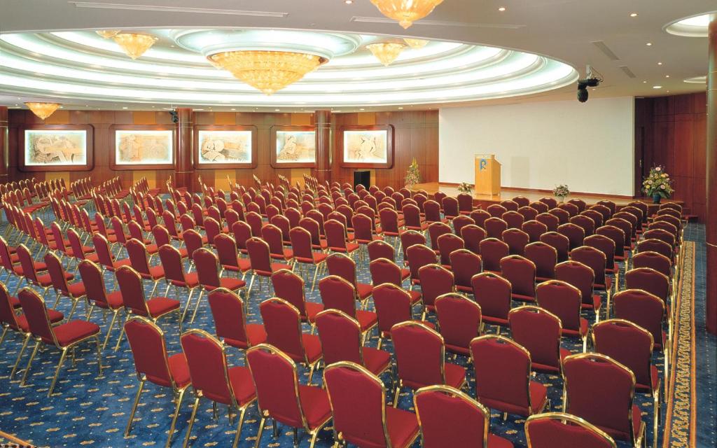 Konferenču zāle ar sarkaniem krēsliem, izkārtotiem rindās, un plašu ekrānu priekšpusē. Telpā ir gaismas un mākslas darbi uz sienām, radot profesionālu atmosfēru.