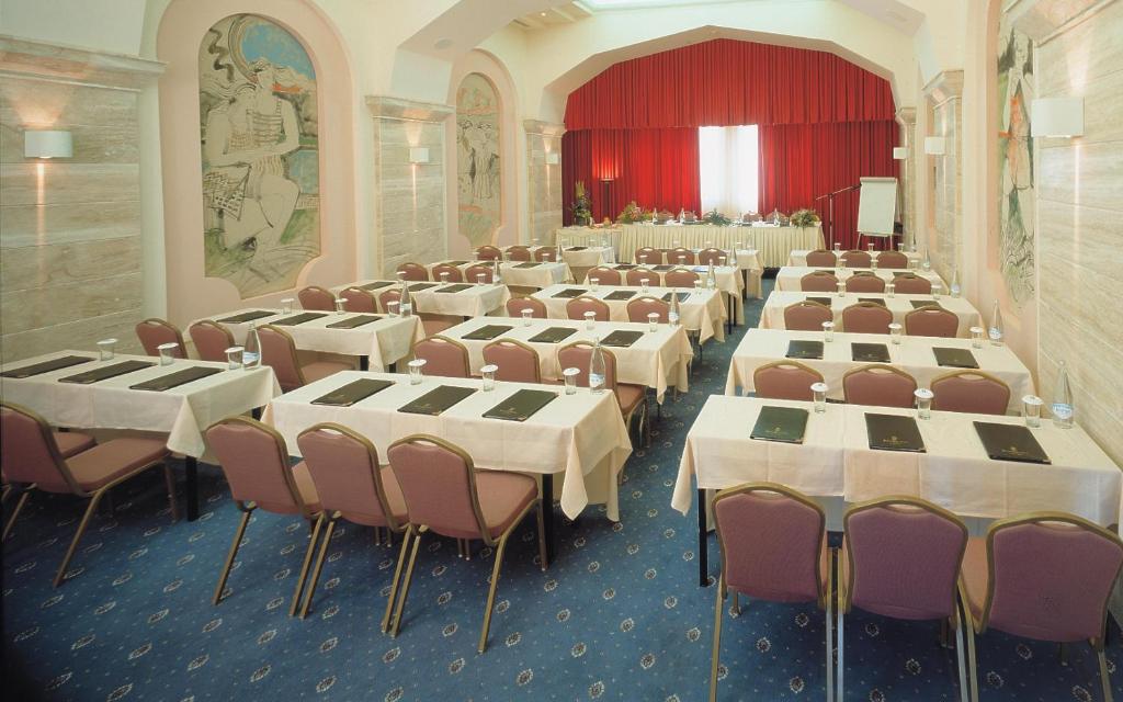 Konferenču zāle ar galdiem un krēsliem, sagatavota sanāksmēm. Telpā ir sarkans aizkars un mākslinieciskas gleznas uz sienām.