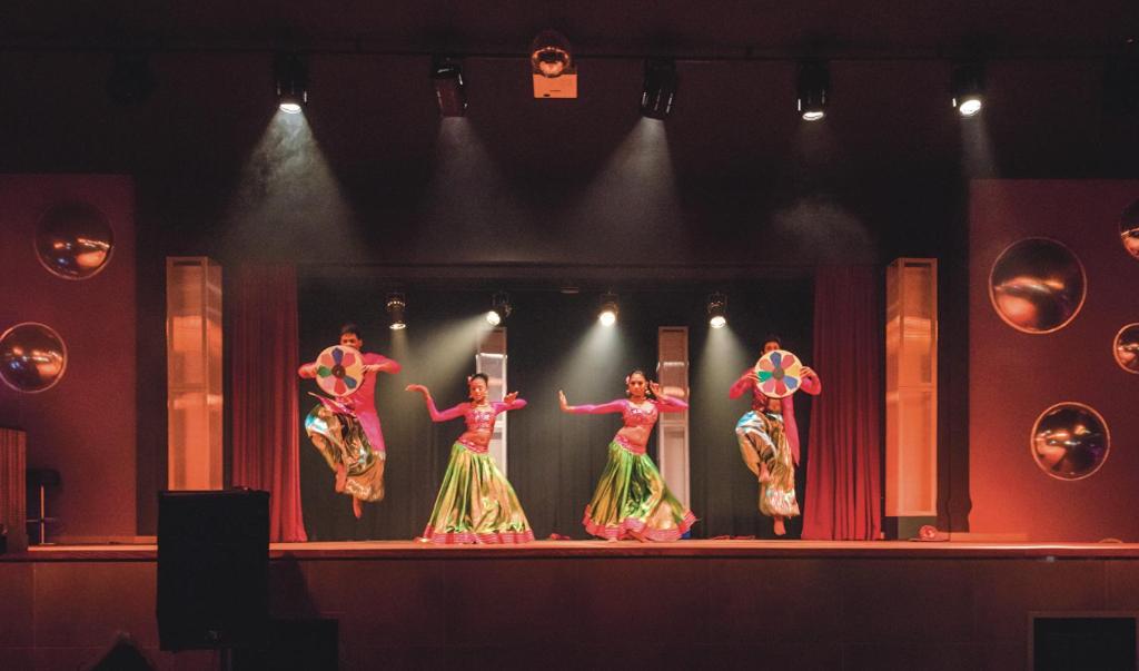 Dejotāju grupa izpilda krāsainu deju uz skatuves, ģērbusies tradicionālās drēbēs ar spīdīgām svārkiem un rotājumiem, kamēr uz skatuves ir gaismas efekti un fonā redzamas aizkars.