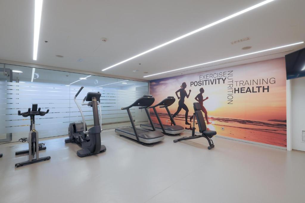 Fitnesa zāle ar modernām trenažieru iekārtām, uzsverot vingrošanas un veselīga dzīvesveida nozīmi. Uz sienas redzams iedvesmojošs uzraksts par veselību un uzturu.