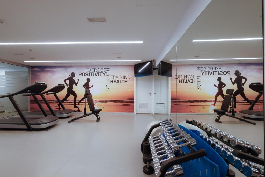 Mūsdienīga sporta zāle ar skrejceliņiem un trenažieriem, uz sienas redzama motivējoša grafika par veselību un vingrošanu.