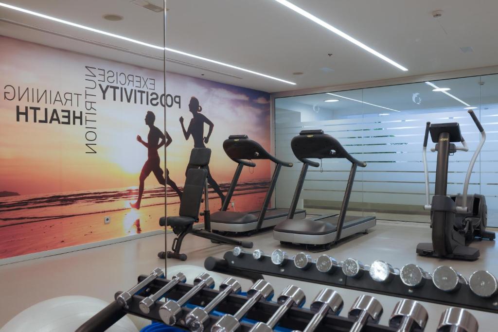 Sporta zāle ar modernām ierīcēm un motivējošu sienas apdari, kas aicina uz aktīvu dzīvesveidu un veselību. Tiek attēloti skriešanas trenažieri un svaru komplekts.