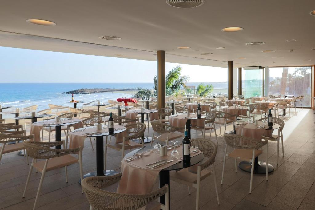 Pludmales restorāna interjers ar skatu uz jūru, kur galds klāts ar baltām galdautām un galda piederumiem. Mūsdienīga atmosfēra ar daudzām vietām un lieliem logiem, caur kuriem ieplūst dabiska gaisma.