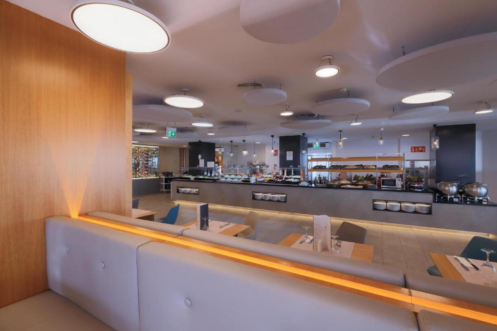 Mūsdienīgs restorāna interjers ar gaišu apgaismojumu un elegantu dizainu, kurā redzama bufete ar dažādiem ēdieniem un galdiņi ar galda piederumiem.