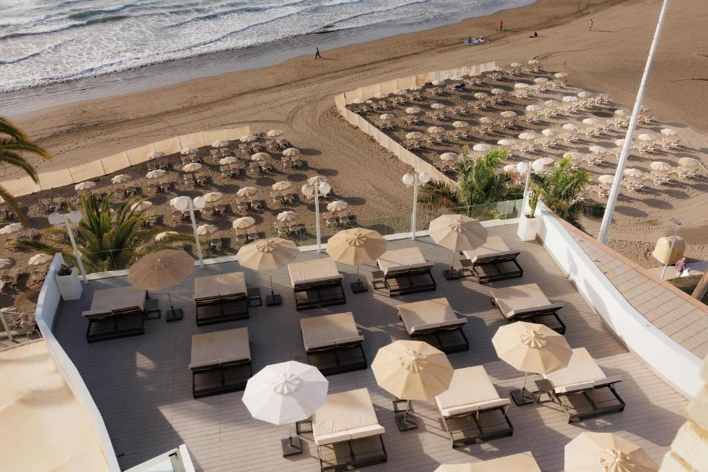 Pludmales skats ar sauļošanās krēsliem un lietussargiem, kas izvietoti piekrastē, blakus viļņiem un smiltīm. Ideāla vieta atpūtai un relaksācijai.