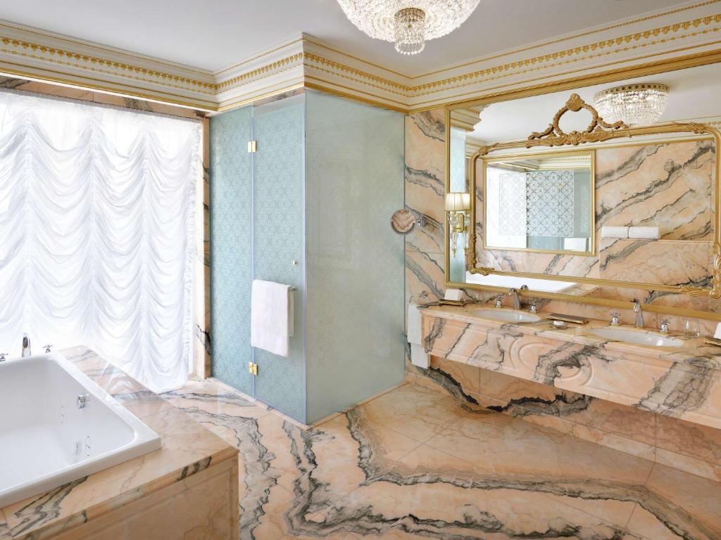 Elegants vannas istabas interjers ar marmora virsmām, stikla dušu un greznu spoguli. Gaisma plūst caur aizkariem, radot relaksējošu atmosfēru.