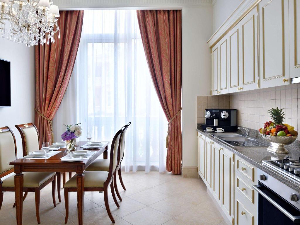 Grezns virtuves un ēdamistabas interjers ar elegantām mēbelēm, lustru un lieliem logiem, kas nodrošina dabīgu apgaismojumu. Uz galda ir klāts galdiņš ar traukiem, bet virtuvē redzami moderni elektroaparāti un augļu kompozīcija.