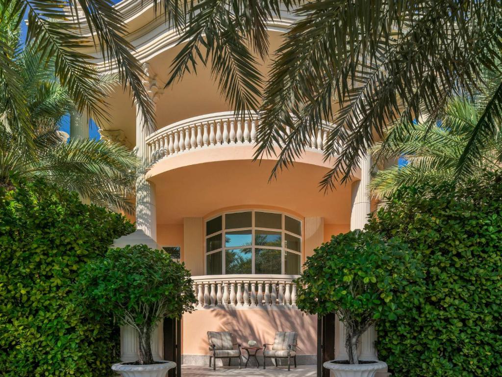 Eleganta mājas ieeja ar balkonu, apjozta ar zaļiem palmu kokiem un krūmiem. Sēdvietas priekšā aicina uz relaksāciju.