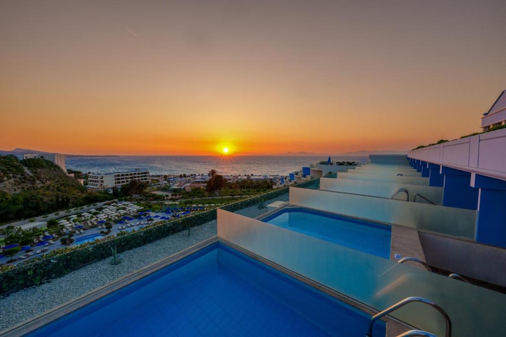 Saulriets pār jūru, redzams no terases ar peldbaseiniem. Skats uz viesnīcu kompleksu un pludmali, kas ir apzaļumota ar sauļošanās krēsliem.