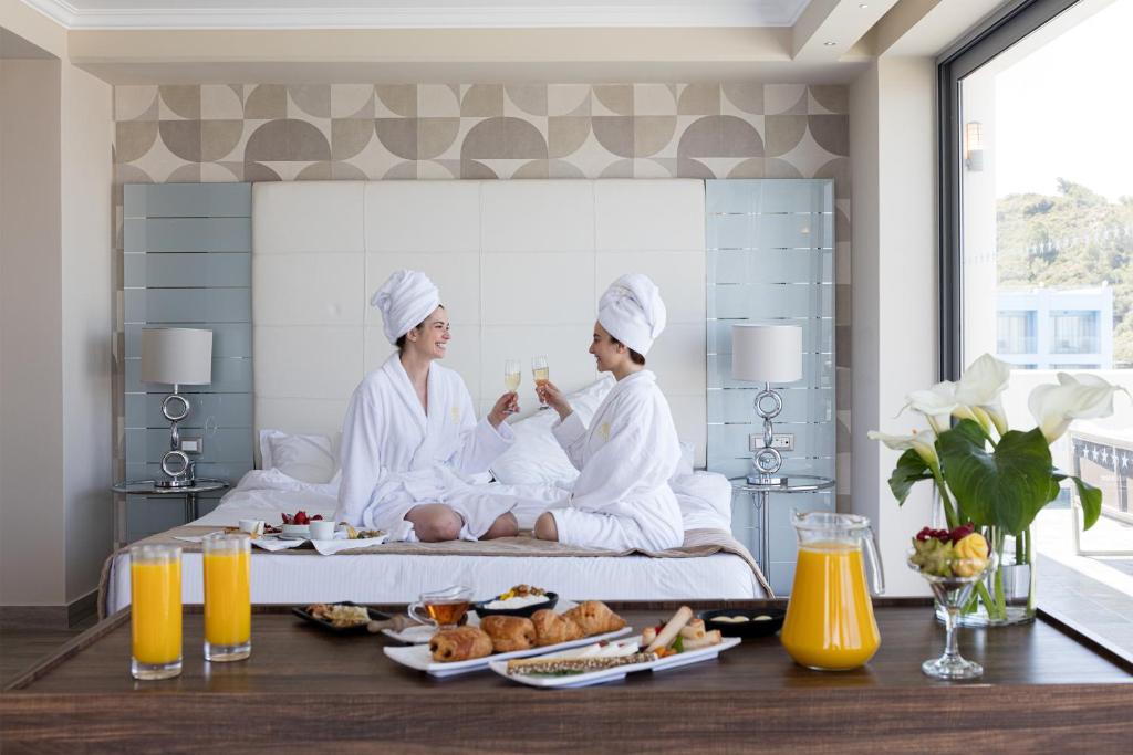 Divas personas, tērptas baltos halātos un ar dvieļiem uz galvas, sēž gultā un svin ar dzērieniem. Uz galda ir brokastu ēdieni un dzērieni, radot relaksējošu un luksusa atmosfēru.