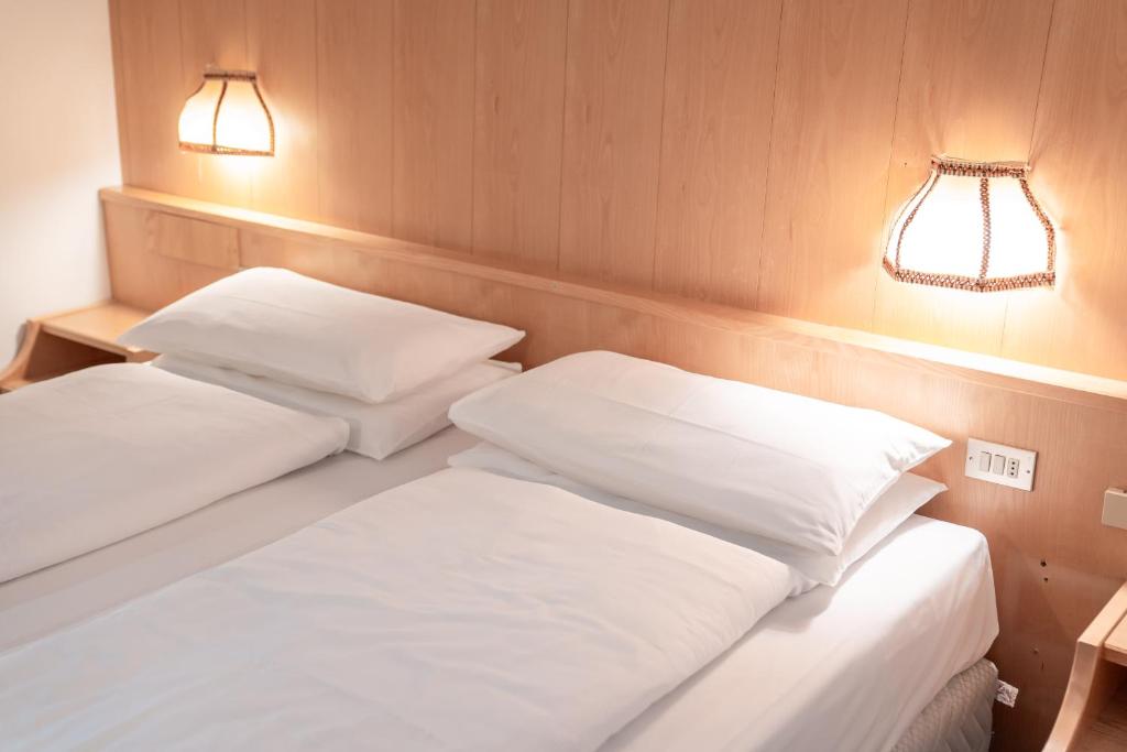 Gultas ar baltām gultasveļām un spilveniem, kas novietotas uz koka gultas. Sienas apgaismojums rada siltu atmosfēru.