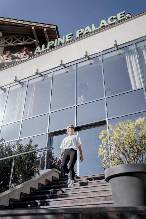 Jauna sieviete, tērpusies brīvā stilā, nolaidjas pa kāpnēm pie modernas viesnīcas ar nosaukumu "Alpine Palace". Viesnīca ir moderna ar lieliem logiem un zaļumiem ap to.