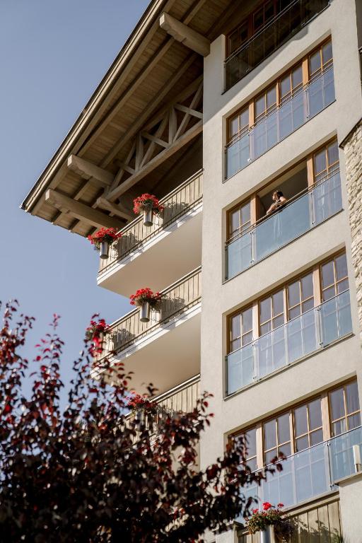 Mūsdienīga ēka ar balkoniem, kuros ziedi rada krāsainu akcentu, un cilvēks, kurš stāv uz balkona. Fons ir skaidrs debesis un koki ar sarkaniem lapu akcentiem.