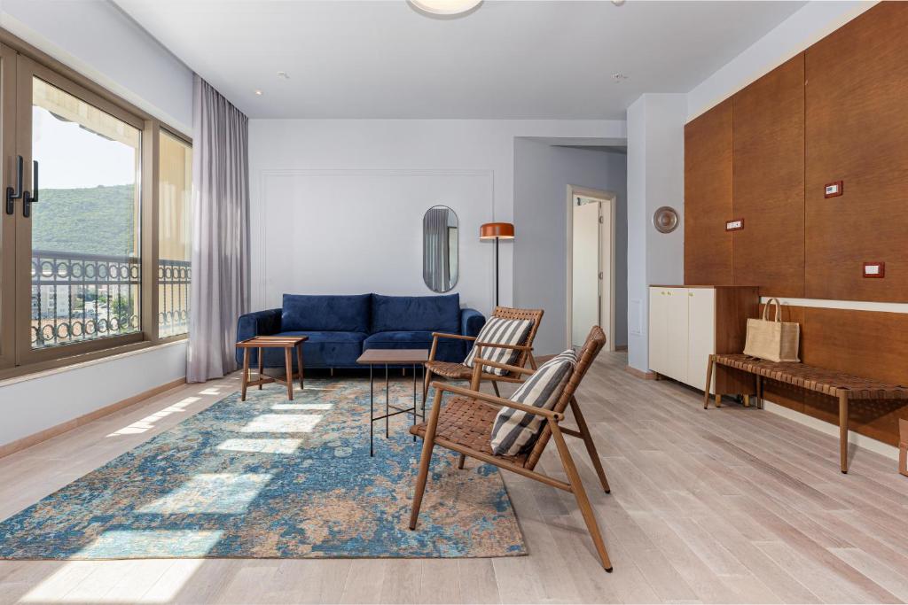 Mūsdienīgs viesistabas interjers ar zilu dīvānu, koka krēsliem un zilu paklāju, kas rada siltu un mājīgu atmosfēru. Gaismas plūdums no logiem uzsver telpas dizainu.