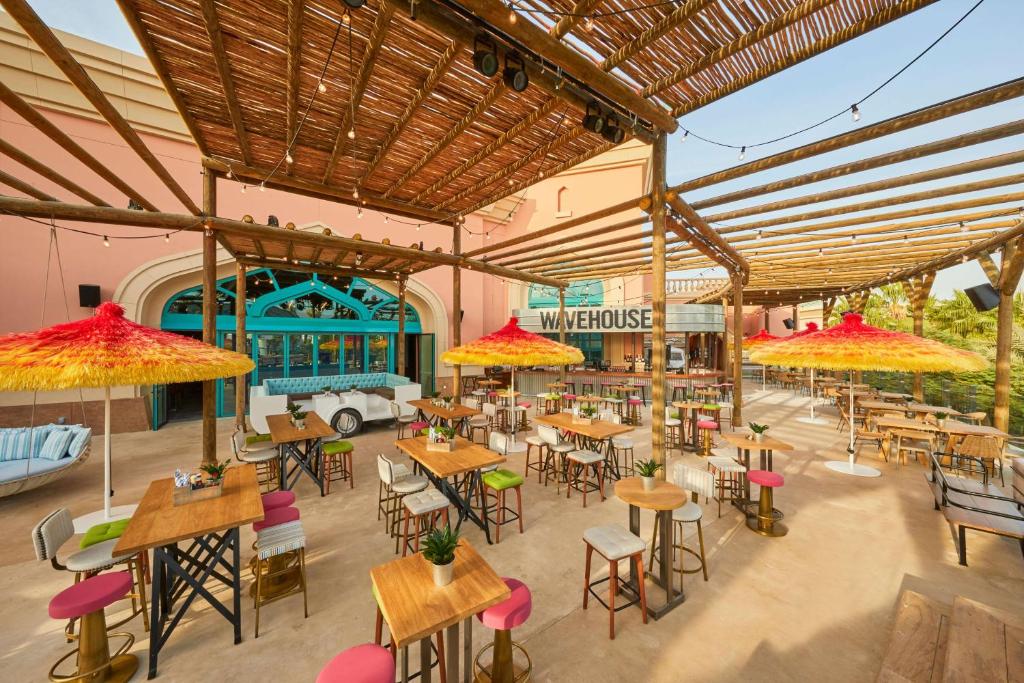 Āra terase ar galdiem un krēsliem, dekorēta ar spilgtiem saules aizsargiem, piedāvā relaksējošu atmosfēru. Fons ietver krāsainas durvis un modernas mēbeles.